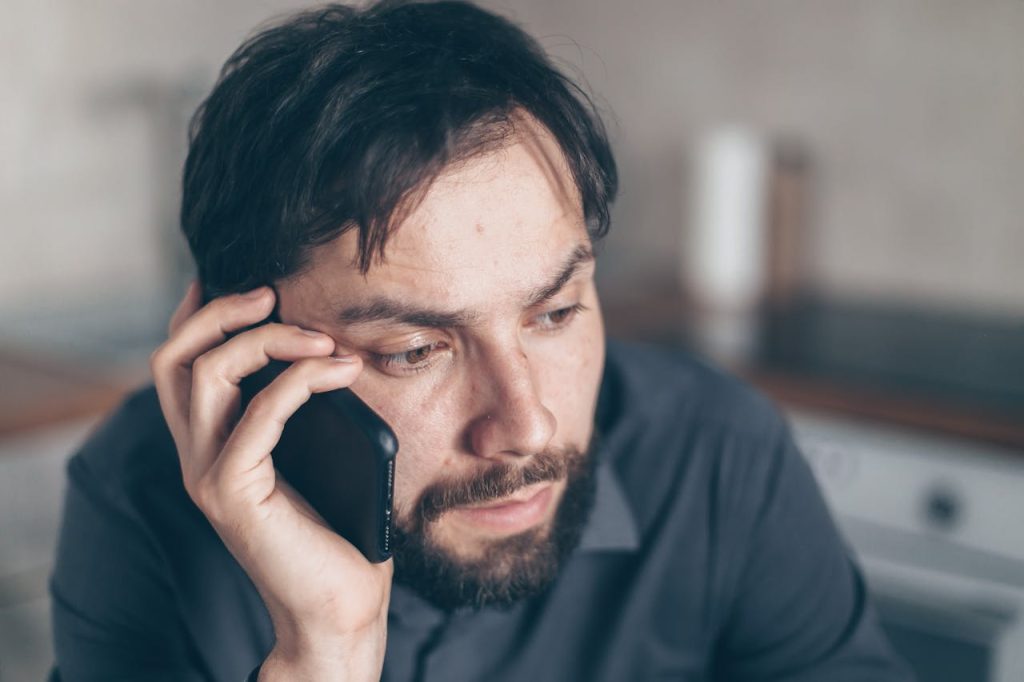 pexels photo 7534387 Thoughtful bearded man using smartphone, sitting indoors with a concerned expression.