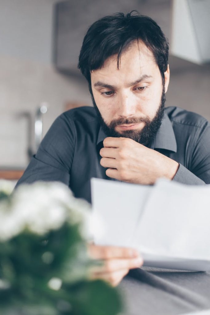 pexels photo 7534780 A bearded man in a gray shirt intently reads documents while seated indoors, appearing concerned.